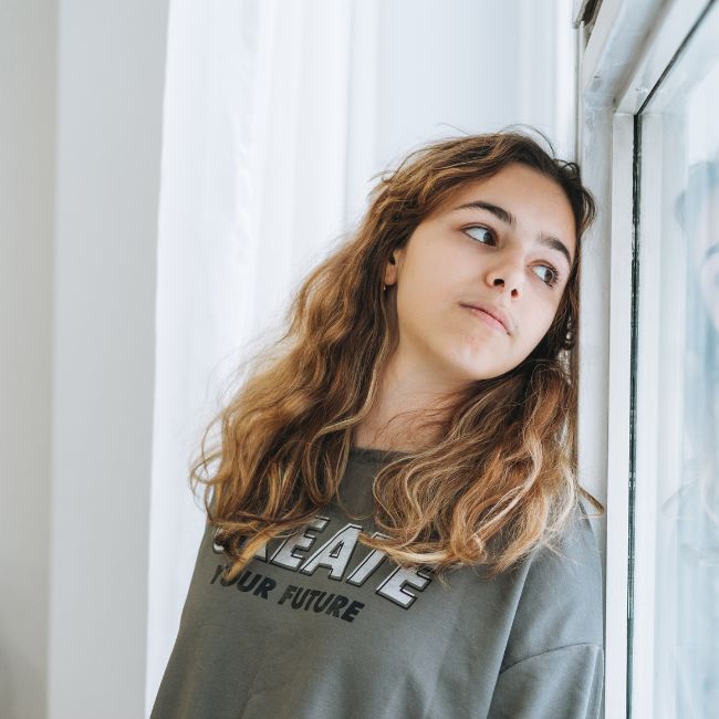 autism in girls, a girl leaning against a window with a grey shirt and brown hair